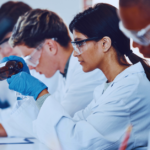 A woman in a white lab coat and blue rubber globes is concentrating while working in a chemistry lab